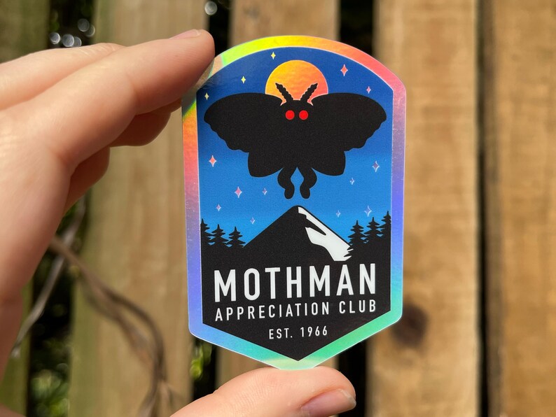 May include: A holographic sticker featuring a black silhouette of a moth with red eyes against a blue night sky with a mountain and trees. The text reads "Mothman Appreciation Club Est. 1966".