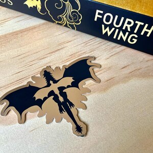 Puede incluir: Una pegatina met&aacute;lica de drag&oacute;n con alas negras y cuerpo dorado, sobre una superficie de madera clara. La pegatina est&aacute; junto a un libro con el t&iacute;tulo "Fourth Wing" en letras doradas sobre fondo azul marino.