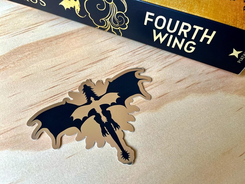 Puede incluir: Una pegatina met&aacute;lica de drag&oacute;n con alas negras y cuerpo dorado, sobre una superficie de madera clara. La pegatina est&aacute; junto a un libro con el t&iacute;tulo "Fourth Wing" en letras doradas sobre fondo azul marino.