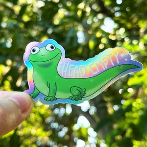 Puede incluir: Una pegatina de lagarto verde de dibujos animados con las palabras "HEAD EMPTY" en texto blanco. La pegatina tiene un borde holográfico de color arcoíris y se sostiene sobre un fondo verde borroso.