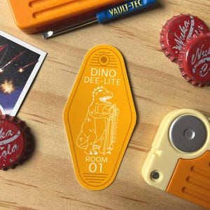 May include: An assortment of Fallout-themed items, including a yellow Dino Dee-Lite room key tag, Nuka Cola bottle caps, and a Vault-Tec tool. The key tag features a dinosaur illustration and the text "DINO DEE-LITE ROOM 01".