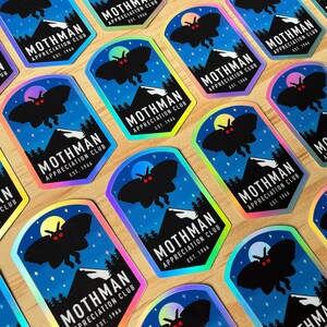 May include: A collection of stickers featuring a black moth silhouette with red eyes, a mountain range, and a full moon against a blue night sky. The text reads "Mothman Appreciation Club Est. 1966". The stickers have a holographic finish.
