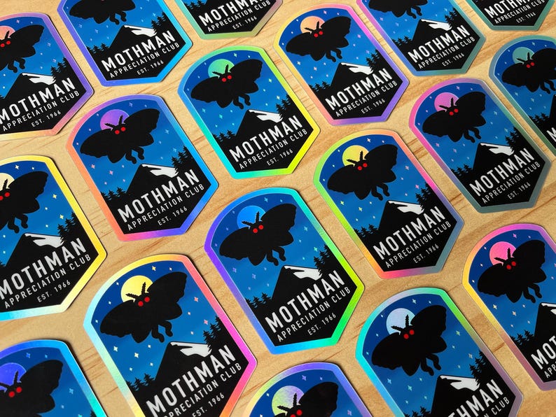 May include: A collection of stickers featuring a black moth silhouette with red eyes, a mountain range, and a full moon against a blue night sky. The text reads "Mothman Appreciation Club Est. 1966". The stickers have a holographic finish.