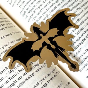 Puede incluir: Un marcap&aacute;ginas con forma de drag&oacute;n dorado y negro. El marcap&aacute;ginas est&aacute; hecho de un material dorado con una silueta de drag&oacute;n negro. El marcap&aacute;ginas est&aacute; sobre las p&aacute;ginas de un libro.