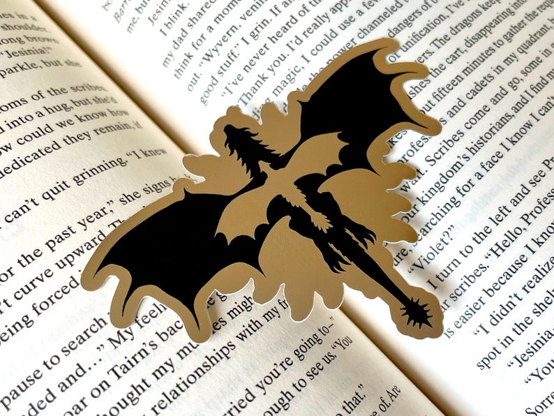Puede incluir: Un marcap&aacute;ginas con forma de drag&oacute;n dorado y negro. El marcap&aacute;ginas est&aacute; hecho de un material dorado con una silueta de drag&oacute;n negro. El marcap&aacute;ginas est&aacute; sobre las p&aacute;ginas de un libro.