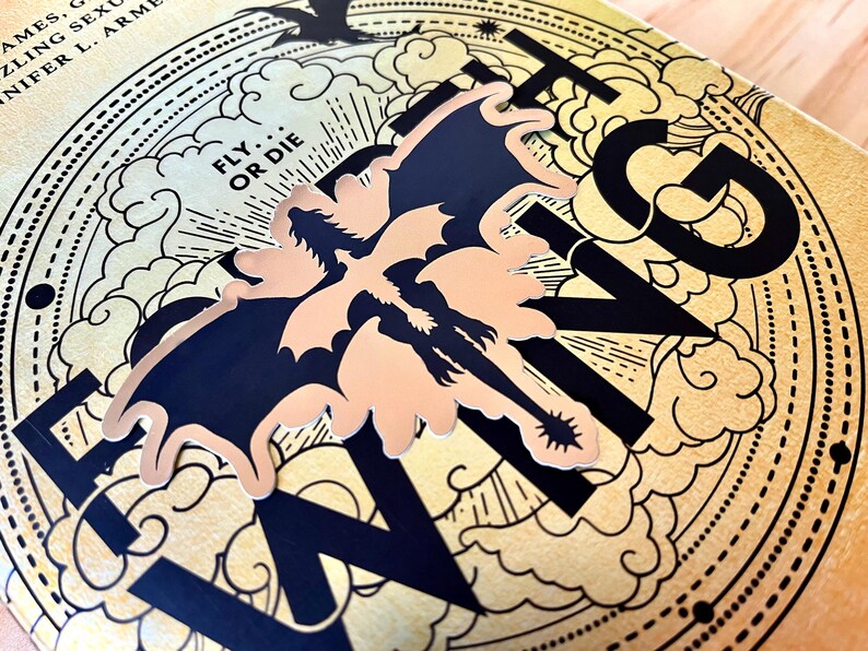 Puede incluir: Primer plano de una portada de libro con la palabra "WING" en letras negras grandes. Un gr&aacute;fico de drag&oacute;n en negro y melocot&oacute;n est&aacute; centrado en la portada. El fondo es dorado con un dise&ntilde;o de nubes y l&iacute;neas. El texto "FLY OR DIE" tambi&eacute;n es visible.