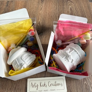 May include: Two white ceramic paint-your-own kits in boxes. One kit features a house-shaped container and the other a carousel. Each box includes paint, brushes, and colourful tissue paper.
