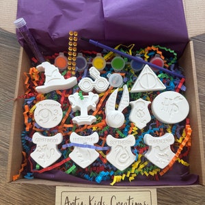 May include: A Harry Potter themed craft kit featuring white ceramic ornaments, paint, brushes, and glitter. The ornaments include the Hogwarts houses, Platform 9 3/4, and other Harry Potter symbols. The kit is packaged in a cardboard box with a purple tissue paper lining.