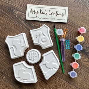 May include: A craft kit featuring white shield-shaped objects with embossed designs, such as a snake, lion, and eagle. The kit includes paint pots, a paintbrush, glitter, and gems. A wooden sign reads "Arty Kids Creations: PLAY - PAINT - CREATE".