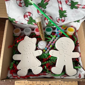 May include: A Christmas craft kit containing two unpainted gingerbread figures, paint pots, glitter, and gems. The kit includes a green paintbrush and is packaged in a box with candy cane-printed tissue paper and red and green paper shreds. The text "Arty Kids Creations" is visible.