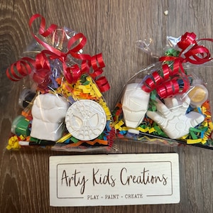 May include: Two clear plastic bags filled with colourful tissue paper, paint, and white ceramic figurines of Spider-Man and Iron Man. The bags are tied with red ribbon and the text "Artsy Kids Creations - PLAY - PAINT - CREATE" is on a wooden sign below.