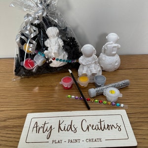 May include: A craft kit featuring two white astronaut figurines, paint, glitter, and rhinestones. The kit is packaged in a clear plastic bag filled with black shredded paper. The bag is tied with a silver ribbon. The kit also includes a wooden sign that reads "Artsy Kids Creations - Play - Paint - Create".