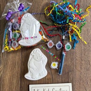 May include: A craft kit featuring two white ceramic monster figurines, paint, glitter, and rhinestones. The kit is packaged in a clear plastic bag with colourful tissue paper. The bag is tied with a purple ribbon. The kit is labelled "Arty Kids Creations - Play - Paint - Create".