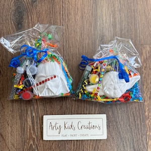 May include: Two cellophane gift bags tied with blue ribbons, each containing a white ceramic figure, paint, and glitter. One bag features a figure with a red beaded necklace, the other a Yoda figure. The bags sit on a wooden surface next to a sign that reads "Arty Kids Creations".