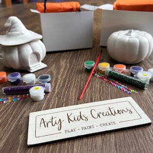 May include: A craft kit featuring two white ceramic pumpkins, one with a witch's hat, paint pots, glitter, and a paintbrush. The kit is labelled "Arty Kids Creations - Play - Paint - Create".