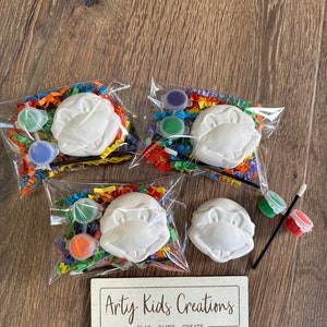 May include: Four paint your own turtle kits with paint, brushes, and colourful tissue paper. The kits are packaged in clear plastic bags. A sign reads "Arty Kids Creations - Play - Paint - Create".