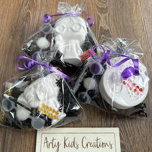 May include: Three white ceramic Halloween-themed paint-your-own kits, each in a clear plastic bag with purple ribbon, black and silver shredded paper, and paint pots. The kits include a skull, a ghost, and a pumpkin. The bags are labelled "Artsy Kids Creations - PLAY - PAINT - CREATE".
