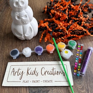 May include: A white ceramic pumpkin stack with three pumpkins, each with a different jack-o-lantern face. The stack is surrounded by craft supplies, including paint, glitter, and rhinestones. The sign reads "Arty Kids Creations - Play - Paint - Create".