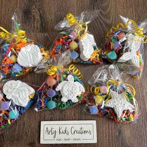 May include: Six clear plastic bags filled with colourful tissue paper and small paint pots. Each bag contains a white ceramic ornament shaped like a sea creature, including a starfish, a turtle, a seashell, and a fish.