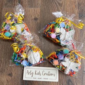 May include: Four cellophane bags containing colourful shredded paper, paint pots, and white ceramic insect figurines. Each bag is tied with a yellow ribbon. A sign reads "Arty Kids Creations PLAY - PAINT - CREATE".