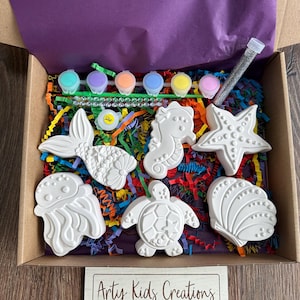 May include: A craft kit containing six white ceramic sea creature figurines, including a mermaid, seahorse, starfish, jellyfish, turtle, and seashell. The kit also includes six small paint pots, two paintbrushes, and a container of glitter. The kit is packaged in a brown cardboard box with a purple tissue paper liner and colourful shredded paper. The box has a label that reads "Arty Kids Creations - PLAY - PAINT - CREATE".