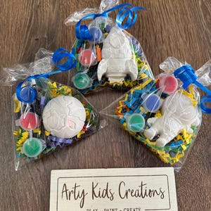 May include: Three clear plastic bags filled with craft supplies for a paint-your-own project. Each bag contains a white ceramic rocket, planet, or astronaut, paint, and colourful tissue paper. The bags are tied with blue ribbon. A sign at the bottom reads "Arty Kids Creations - Play - Paint - Create."
