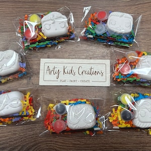 May include: Seven individual packages containing a white ceramic figure, paint pots, a paintbrush, and colourful shredded paper. The packages are labelled "Arty Kids Creations - Play - Paint - Create".