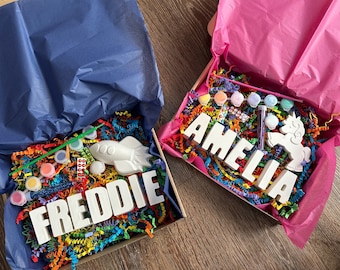 Könnte beinhalten: Zwei Bastelsets für Kinder, eines mit einer Rakete und dem Namen Freddie, das andere mit einem Einhorn und dem Namen Amelia. Beide Sets enthalten Farbe, Glitzer und bunte Papierschnitzel.