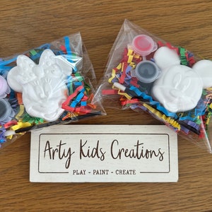 May include: Two paint-your-own ceramic figurines of Mickey Mouse and Minnie Mouse, packaged in clear plastic bags with colourful tissue paper and paint pots. The bags are on a wooden surface with a sign that reads "Arty Kids Creations - PLAY - PAINT - CREATE".