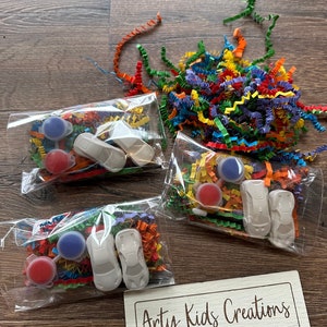 May include: Three clear plastic bags containing paint supplies and toy cars, with colorful shredded paper. Each bag includes two small paint pots, a toy car, and decorative paper. A wooden sign with the text "Arty Kids Creations" is also visible.