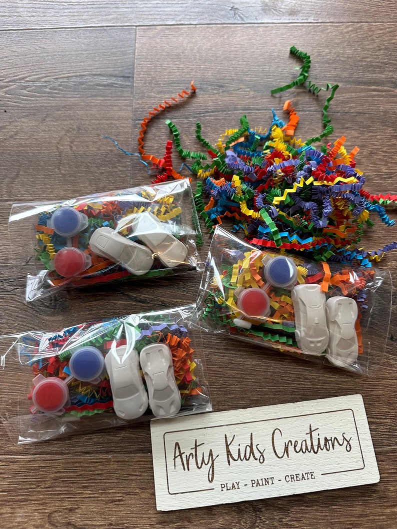 May include: Three clear plastic bags containing paint supplies and toy cars, with colorful shredded paper. Each bag includes two small paint pots, a toy car, and decorative paper. A wooden sign with the text "Arty Kids Creations" is also visible.