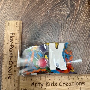 May include: A clear plastic bag filled with colorful confetti, paint pots, and a white wooden letter K. The bag is labeled "Arty Kids Creations".