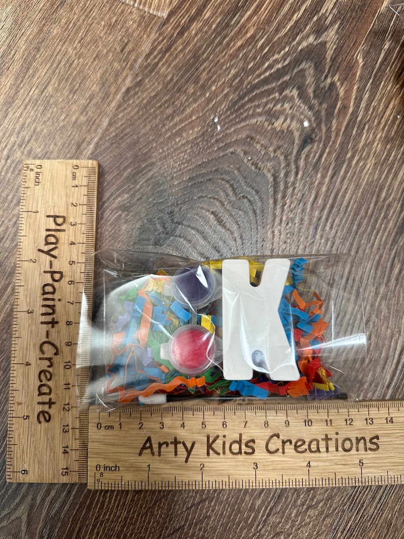 May include: A clear plastic bag filled with colorful confetti, paint pots, and a white wooden letter K. The bag is labeled "Arty Kids Creations".