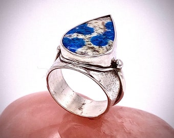 Könnte beinhalten: Ein silberner Ring mit einem tropfenförmigen blauen und weißen Stein, der in der Mitte eingesetzt ist. Der Ring hat eine strukturierte Band und eine kleine silberne Perle an der Seite.
