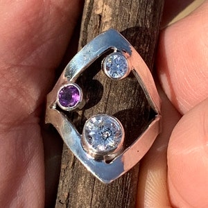 May include: A silver ring with a geometric design featuring three round stones. The stones are clear and purple.
