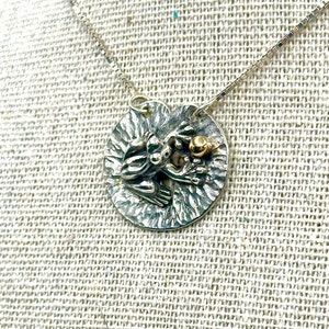 May include: A silver necklace with a round pendant featuring a detailed frog sculpture holding a small gold ball. The pendant has a textured surface.