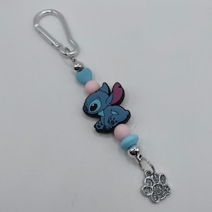 May include: A keychain featuring a blue cartoon character with pink ears and a silver paw print charm. The keychain includes blue and pink beads, a silver clasp, and a silver bead.