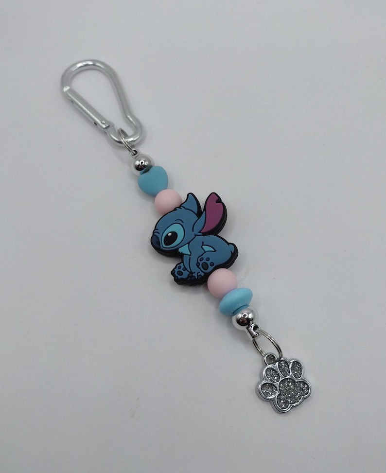May include: A keychain featuring a blue cartoon character with pink ears and a silver paw print charm. The keychain includes blue and pink beads, a silver clasp, and a silver bead.