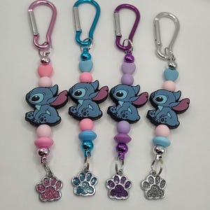 May include: Four keychains with colorful beads, cartoon character charms, and paw print charms. Each keychain has a different colored carabiner clip: pink, blue, purple, and silver. The charms are blue with pink ears.
