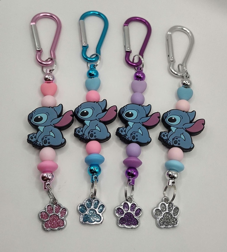May include: Four keychains with colorful beads, cartoon character charms, and paw print charms. Each keychain has a different colored carabiner clip: pink, blue, purple, and silver. The charms are blue with pink ears.