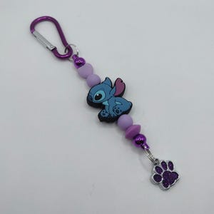 May include: A purple carabiner clip keychain with a blue cartoon character, lavender beads, and a silver charm with a purple glitter paw print. The keychain is designed with a series of purple beads.
