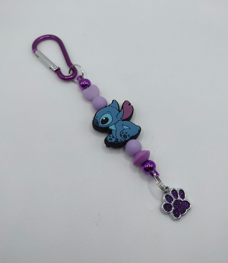 May include: A purple carabiner clip keychain with a blue cartoon character, lavender beads, and a silver charm with a purple glitter paw print. The keychain is designed with a series of purple beads.