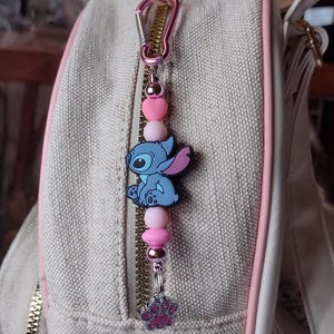 May include: A pink and silver charm with a blue Stitch character, pink heart and round beads, and a paw print charm. The charm is attached to a pink carabiner clip, hanging from a beige bag with a pink zipper.