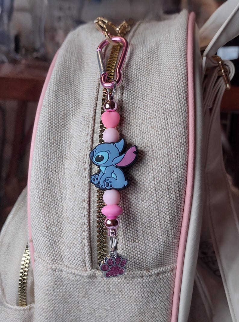 May include: A pink and silver charm with a blue Stitch character, pink heart and round beads, and a paw print charm. The charm is attached to a pink carabiner clip, hanging from a beige bag with a pink zipper.
