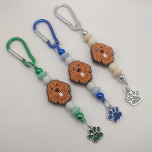May include: Three dog-themed keychains featuring a brown doodle dog charm, colourful beads, and a paw print charm. Each keychain has a different coloured carabiner clip: green, blue, and silver. These keychains are designed for pet lovers.