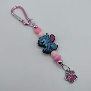 May include: A pink keychain featuring a blue cartoon character, pink beads, and a silver paw print charm. The keychain has a pink carabiner clip for easy attachment to bags or keys. The charm is a cute accessory.