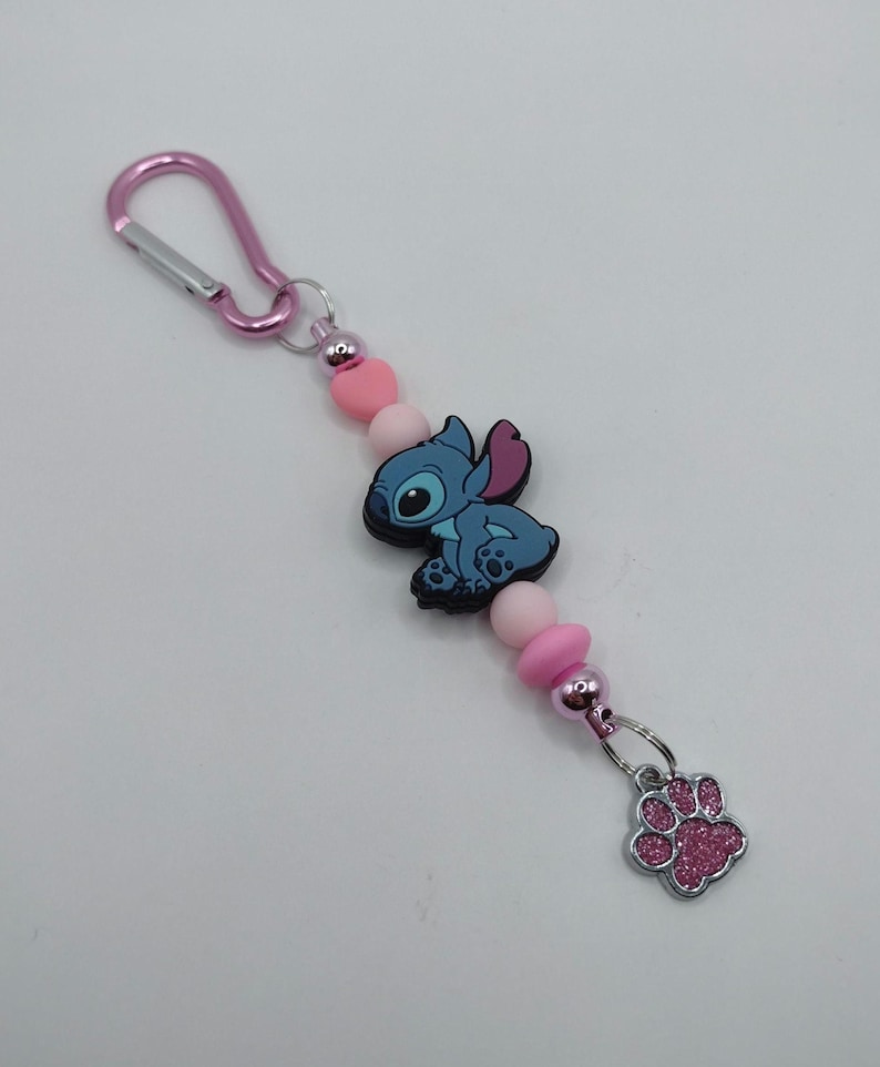 May include: A pink keychain featuring a blue cartoon character, pink beads, and a silver paw print charm. The keychain has a pink carabiner clip for easy attachment to bags or keys. The charm is a cute accessory.