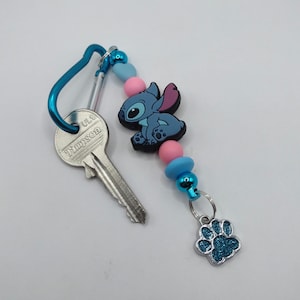 May include: A blue carabiner clip with a key and a Stitch-themed keychain. The keychain features a blue Stitch figure, pink and blue beads, and a glittery blue paw print charm. The key has the word "Timpson" printed on it.