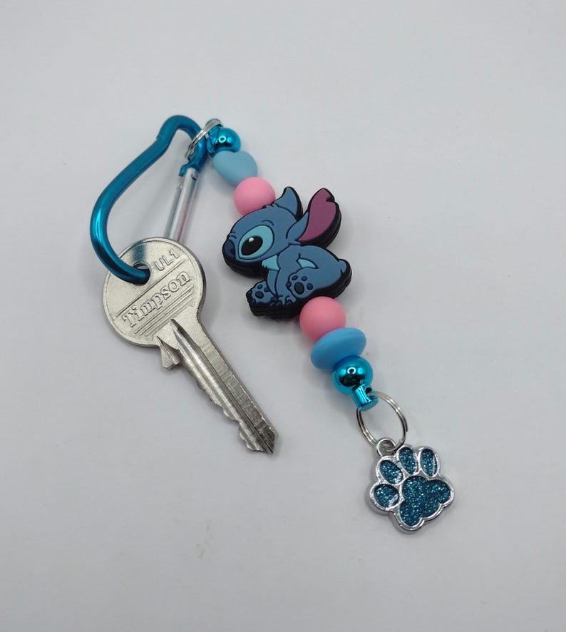 May include: A blue carabiner clip with a key and a Stitch-themed keychain. The keychain features a blue Stitch figure, pink and blue beads, and a glittery blue paw print charm. The key has the word "Timpson" printed on it.
