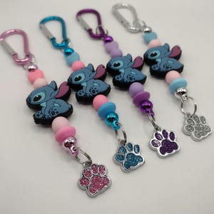 May include: Keychains featuring a blue cartoon character with large ears and a paw print charm. The keychains have colorful beads in pink, blue, and purple, and silver-tone carabiner clips.